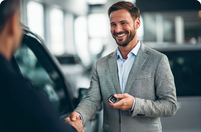 A person wearing overalls handing over a car key to someone. There is a car in the background.