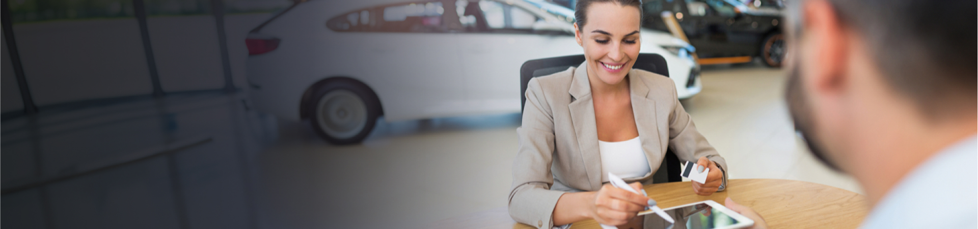 A woman signing something on an ipad whilst holding a bank card. There is a man sat across the desk from her and a car in the background. 